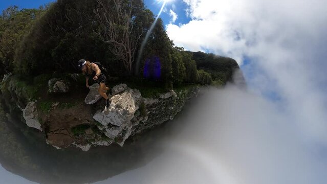A young and strong solo hiker is walking along the steep edge of Espigao Amorelo in Madeira with thin clouds around him as he takes a dangerous step up to a rock on the cliffside.