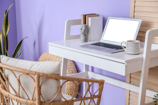 Workplace with modern laptop, cup and books near violet wall in room
