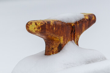 An old rusty yellow colored metal mooring for large industrial boats on a wooden wharf covered in fresh white snow.  The ballard is an industrial style and is covered in brown rust. 