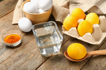 Homemade dyed Easter eggs, bowl with water and turmeric powder on wooden table, closeup