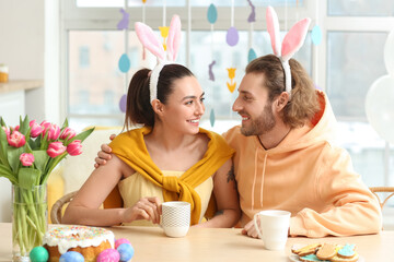 Happy couple in bunny ears with cups of cocoa sitting at table on Easter day