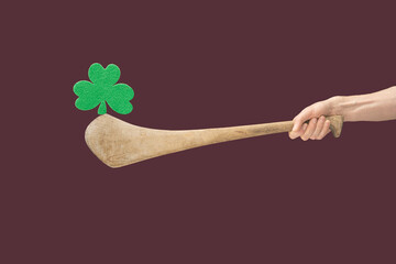 Man hands hold hurly sticks – hurleys. Green clover balancing  on top of it against a green background. Ireland traditional sport minimal concept. Plum background - obrazy, fototapety, plakaty