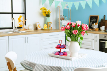 Stand with Easter eggs and vase with tulips on dining table in kitchen interior