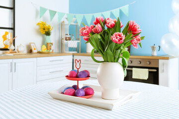 Stand with Easter eggs and vase with tulips on dining table in kitchen