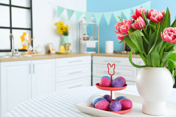 Stand with Easter eggs and vase with tulips on dining table in kitchen