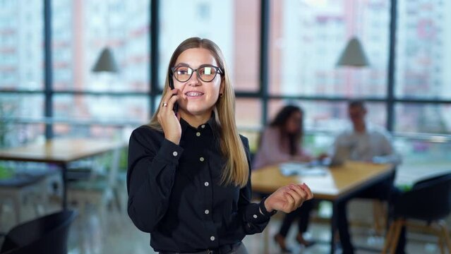 Pretty long-haired woman in glasses chats on the phone. Smiling lady having phone conversation in big spacious office. Colleagues sit at the desk at backdrop in blur.
