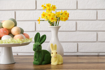 Stand with Easter eggs, rabbit and narcissus flowers on wooden counter near white brick wall