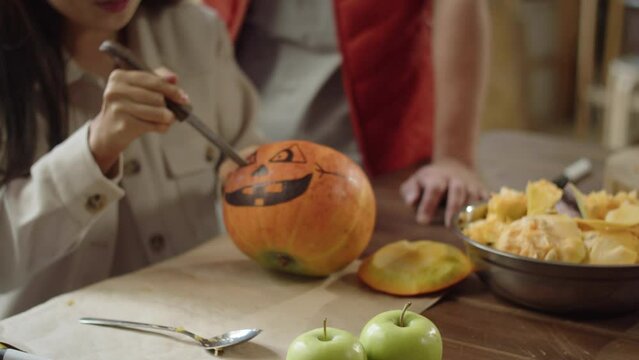 An adult girl is carving eyes for a halloween pumpkin while her friend oversees the process