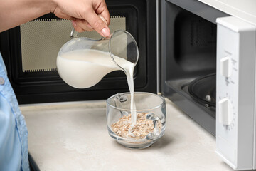 Woman adding milk to oatmeal on table near opened microwave oven, closeup