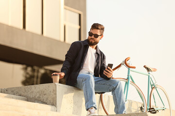 Fototapeta na wymiar Stylish bearded man with mobile phone, cup of coffee and bicycle in city