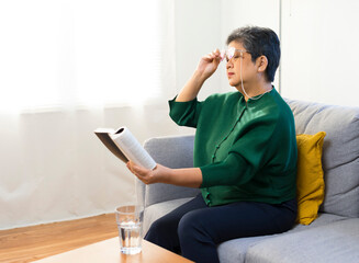Elderly woman having troubles with long eye-sight trying to read a book, sitting on sofa at home.