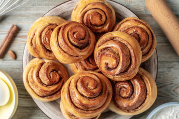 There is a plate with homemade fresh delicious cinnamon rolls, ingredients for their preparation and kitchen tools on the wooden table . Top view