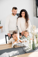 Obraz na płótnie Canvas Joyful family with small son and daughter having a breakfast, spending weekends at home. Beautiful happy parents standing at kitchen and smiling, small kids sitting at the table and eating croissants