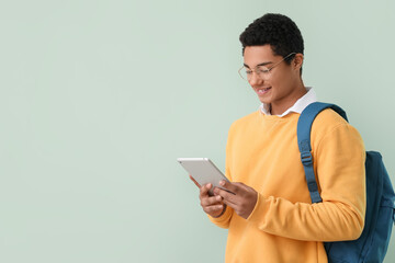 Male African-American student with backpack and tablet computer on color background