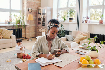 Serious modern young African-American mother in headscarf sitting at table and making notes in...