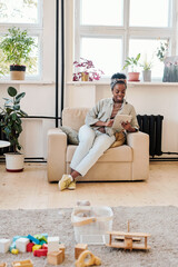 Obraz premium Smiling young African-American mother sitting in armchair and using tablet while reading article about children on internet