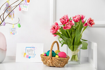 Basket with painted eggs, tulips and greeting card with text HAPPY EASTER on table near white wall