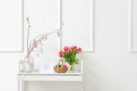 Beautiful Easter composition with basket, painted eggs, flowers and greeting card on table near white wall