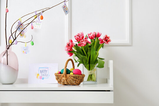 Basket with painted eggs, tulips and greeting card with text HAPPY EASTER on table near white wall