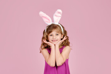 Obraz na płótnie Canvas Adorable little coquette with long curly hair in stylish fuchsia colored dress and bunny ears touching cheeks and smiling at camera, against ink background in studio