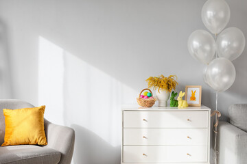 Chest of drawers with basket of Easter eggs, flowers and decor near light wall in room