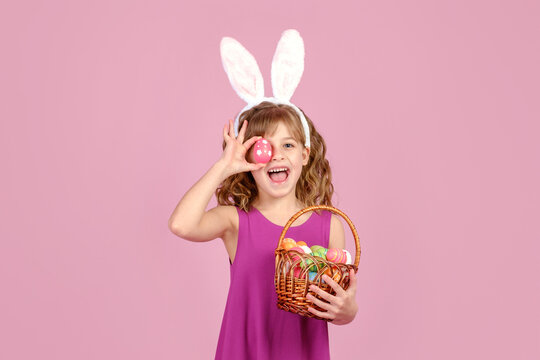 Little beautiful smiling girl in Easter bunny ears holds in hand a basket with Easter eggs, and covering eye with colorful egg on pink background 