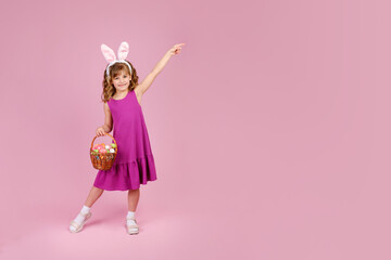 Full body little beautiful smiling girl in Easter bunny ears holds a basket with eggs on pink...