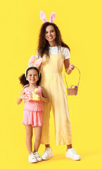 Little African-American girl with Easter rabbits, her mother and eggs on yellow background