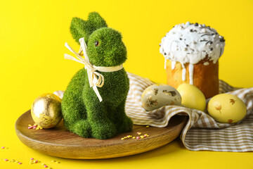 Plate with cute Easter bunny, cake and painted eggs on yellow background