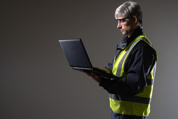 Laptop engineer. Experienced engineer. Gray-haired man in yellow vest. Portrait of engineer on studio background. Man is holding open laptop. Engineering software concept. Builder or architect