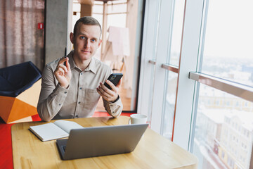 Successful young man is sitting at a table in a cafe-restaurant indoors, working and studying on a laptop computer,holding a mobile phone with a blank screen. Freelancer mobile office business concept