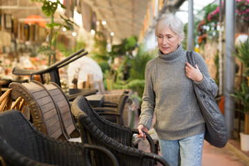 Interested elderly woman visiting furnishing store looking for practical plastic wicker chairs for...