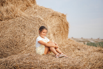 Naklejka na ściany i meble Small female kid with sly facial expression on haystack looking somewhere away slightly smiling. Time away from city in country field with tons of hayricks.