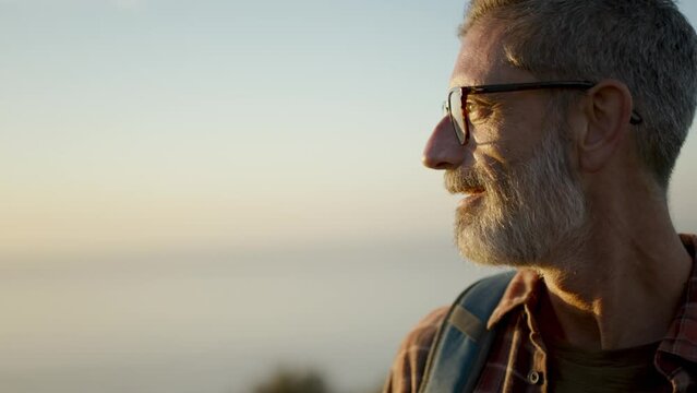 Close up of a senior man wearing backpack on a holiday. Side view of a male hiker standing on a hill and looking at sunset. Panning shot.