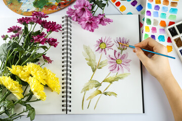 Woman painting chrysanthemums in sketchbook at white table, top view