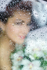 silhouette of a bride holding a bouquet of white flowers, behind a transparent glass covered with water drops