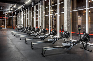Modern rowing machines and exercise bikes lined near huge windows in spacious, empty gym interior....