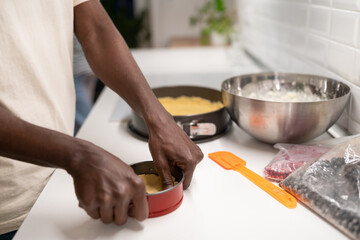 African man making homemade cake for celebrating special occasion, cropped photo. Black guy standing in kitchen putting shortbread pastry in springform pan, selective focus. Cooking, baking concept