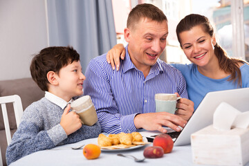 Cheerful family with preteen son spending time together at home, drinking tea with sweets and using laptop