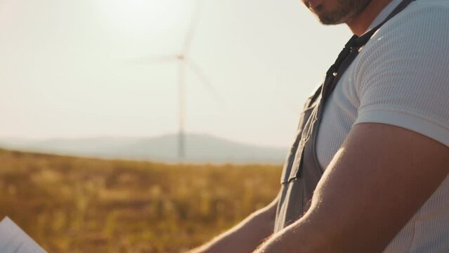Closeup Indian technician in overalls, helmet and glasses checking blueprints of windmills while standing at rural area. Industrial worker controlling process of green energy production