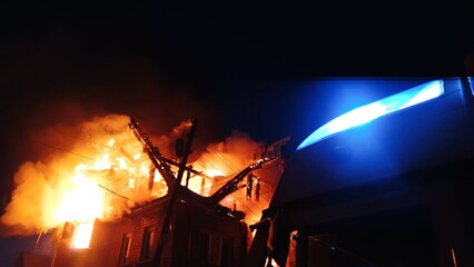 Fototapeta premium The roof of the house is on fire. The residential building burn, village. Firefighters put out a fire. Fire Department putting out flame. Fire truck, headlights and panel, siren, signal, alarm.