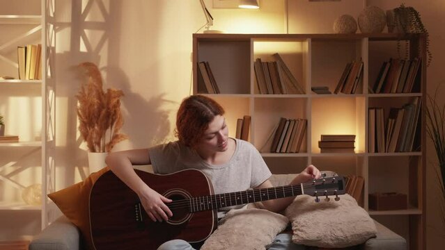 Guitar tuning. Music practice. Quarantine leisure. Woman guitarist adjusting pegs and gears on string acoustic instrument on couch at home living room.