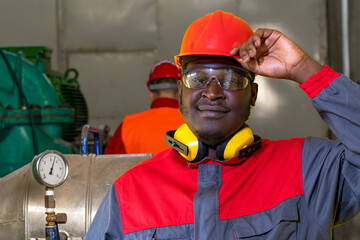 Portrait Of Young African American Worker In Personal Protective Equipment In Industrial Interior. Black Worker In Red Helmet, Safety Goggles, Hearing Protection Equipment And Work Uniform.