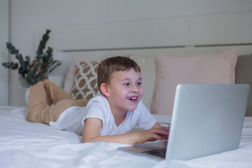 Adorable schoolboy uses internet while lying on bed at home. Online school. Cute 7 year old boy plays video games on a laptop.