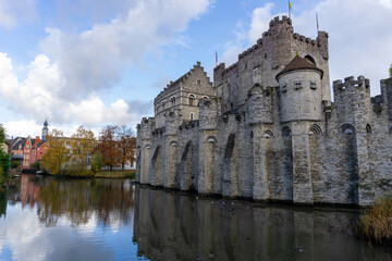 Fototapeta na wymiar Ghent Castle with its reflection in the water on a day with some clouds. Trees at the end of the photo with fall colors.
