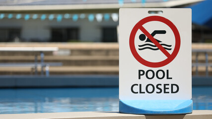 A bright white sign at a local outdoor pool facility saying pool is closed. No entry to water. Clear clean blue water at public baths and no swimming symbol.