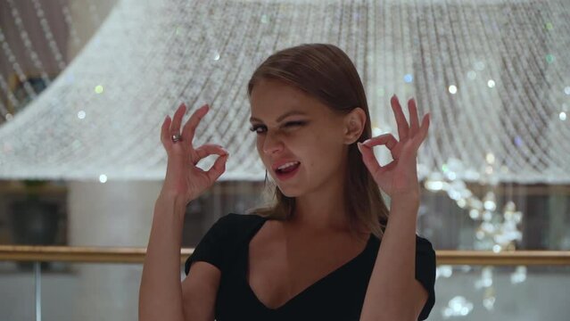 Caucasian girl gestures ok sign. A beautiful European woman in a black dress on a background of a large crystal chandelier. Portrait of a pretty lady.