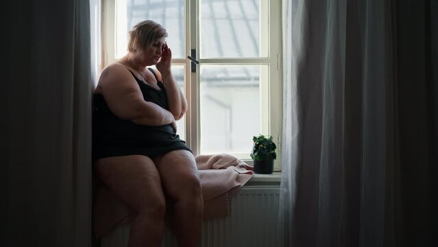 Depressed lonely fat woman sitting aby window at home and thinking.