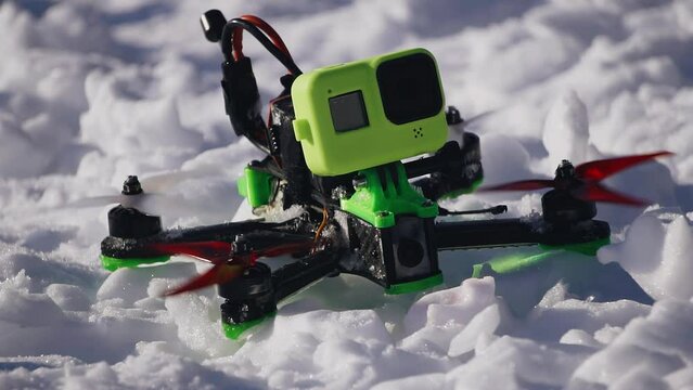 The quadcopter stands on the snow and then starts and takes off into the air. He goes out of the frame up