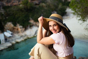 Happy traveller with a hat in front of the beach at sunset. Beautiful woman portrait on a cliff.